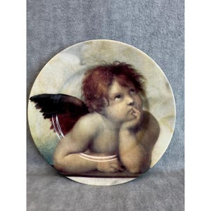 May include: Round decorative plate featuring a Renaissance-style cherub with red hair, a thoughtful expression, and a finger to its lips. The cherub has a dark wing and is set against a cloudy background.