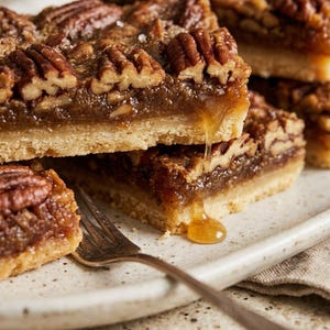 Op de afbeelding: Een stapel vierkante pecannotentaartrepen met een goudbruine zanddeegbodem en een dikke, kleverige karamelvulling met hele pecannoten. Een druppel siroop loopt langs de rand naar beneden. Een zilveren vork ligt ernaast. Tekst luidt MsApronista.