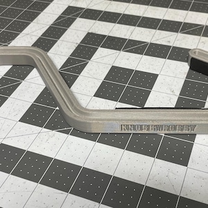 May include: A gray, curved, 3D-printed hook with a black rubber grip. The text "KNOPBYROBBY" is printed on the side. The hook is designed to hang items and is shown on a black and white checkered surface.