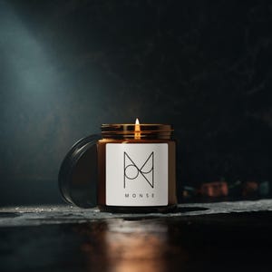 May include: A lit candle in a brown glass jar with a gold lid and a white label. The label features a geometric design and the word "MONSE" in black. The candle sits on a dark surface with the lid open.