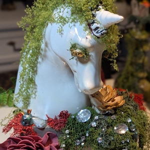 May include: A white ceramic unicorn bust adorned with green moss, red and brown flowers, and clear crystals. The unicorn has a white horn and is set in a wooden box. The bust is decorated with gold accents.