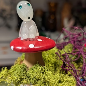 Puede incluir: Una figura caprichosa de una pequeña criatura translúcida con ojos turquesa, posada sobre una seta roja con lunares blancos. La seta se asienta sobre un lecho de musgo verde, creando una estética de cuento de hadas. Un artículo decorativo único.