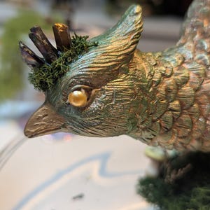 May include: A detailed, close-up of a decorative bird figurine. The bird is primarily green with gold accents, featuring a textured surface. It has a prominent eye, a crown of crystals, and is adorned with moss. The figurine is set against a blurred background.