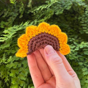 May include: A handmade crochet sunflower with a yellow petal edge and a brown center. The sunflower is held in front of a green leafy background. The crochet flower is a decorative item.