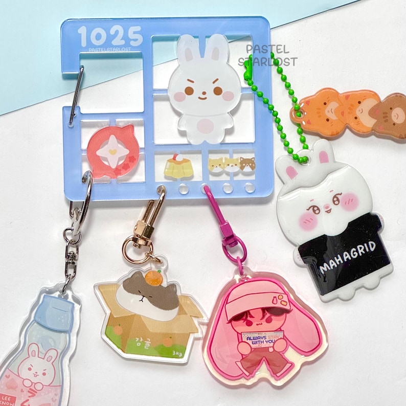 May include: A selection of acrylic keyrings with cartoon characters and food designs. The keyrings include a blue grid with the text "1025" and "PASTEL STARLOST", a white bunny, a pink character holding a sign that says "ALWAYS WITH YOU", and a cat in a box.