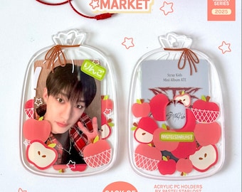 May include: Clear acrylic PC holders shaped like sacks, filled with red apple designs and a photo card. The text "SACK OF APPLES" and "STARLOST MARKET" are visible. The holders are part of the "VOL 1 MARKET SERIES 2025".