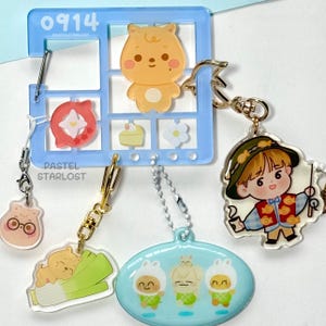 May include: A selection of acrylic keyrings with cartoon characters and food designs. The keyrings include a bear, a boy in a hat, and three figures in bunny costumes. A blue window-shaped holder with the text "0914" and "PASTELSTARLOST" is also visible.