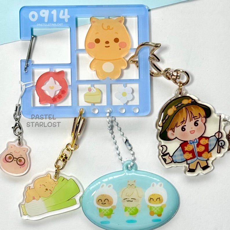 May include: A selection of acrylic keyrings with cartoon characters and food designs. The keyrings include a bear, a boy in a hat, and three figures in bunny costumes. A blue window-shaped holder with the text "0914" and "PASTELSTARLOST" is also visible.
