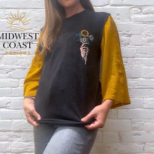 May include: Black t-shirt with mustard yellow sleeves. The shirt features a floral design held by a skeleton hand. The text "MIDWEST COAST DESIGNS" is printed on the left side of the shirt.