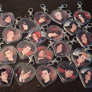 May include: Heart-shaped keychains with various portraits. Each keychain features a different person's portrait, with a silver-colored clasp. The portraits are in a variety of styles and colors, with some featuring text. The keychains are arranged on a dark surface.