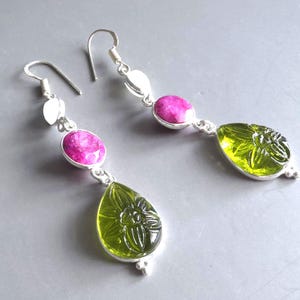 May include: A pair of dangle earrings with a silver finish. Each earring features a pink oval stone, a silver leaf-shaped accent, and a green, teardrop-shaped stone with a floral design. The earrings are designed for pierced ears.