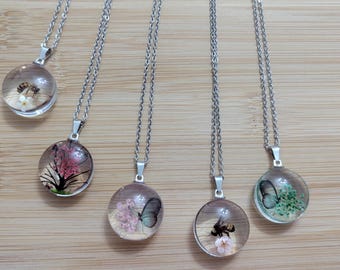 May include: Five silver pendant necklaces with clear, round resin charms. Each charm contains a different natural element, including a bee, flowers, and butterflies. The necklaces are displayed on a light brown wooden surface.