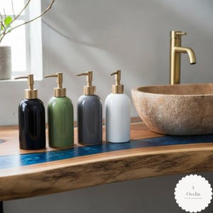 May include: Four ceramic soap dispensers in black, green, grey, and white, each with a gold-coloured pump. The dispensers are on a wooden countertop with a blue resin inlay, near a stone sink and gold tap.