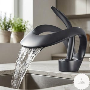 Könnte beinhalten: Ein mattschwarzer Badezimmerwasserhahn mit geschwungenem, skulpturalem Design und einem breiten, offenen Auslauf. Wasser fließt in einem flachen, wasserfallartigen Strahl in ein Edelstahlbecken. Der Wasserhahn hat einen Hebelgriff und den Text Ovelin.