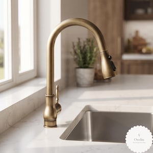 Può includere: Un rubinetto da cucina ad arco alto con finitura in ottone spazzolato, dotato di una doccetta estraibile con interruttore nero e una leva laterale. Il rubinetto è posizionato accanto a un lavello in acciaio inossidabile su un piano in marmo.