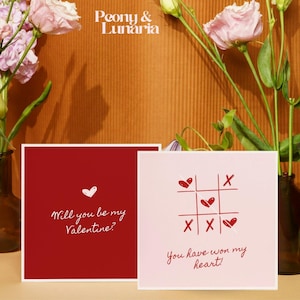 Könnte beinhalten: Zwei quadratische Valentinstagskarten. Eine ist rot mit dem Text "Willst du mein Valentin sein?" und einem weißen Herzen. Die andere ist rosa mit einem Tic-Tac-Toe-Spiel mit Herzen und dem Text "Du hast mein Herz gewonnen!" Die Karten liegen neben rosa Blumen.
