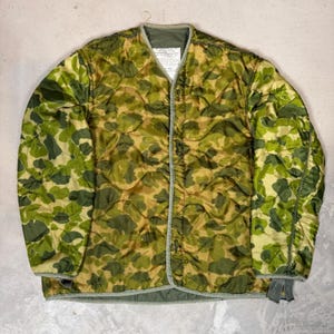 May include: A quilted liner jacket featuring a camouflage pattern in shades of olive green, brown, and tan. The jacket has a V-neckline, long sleeves, and a front button closure with olive green trim. A white rectangular label is visible at the neck.