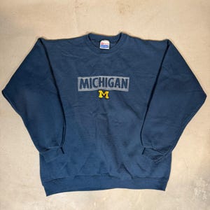Peut inclure: Un sweat-shirt bleu marine à col rond avec le mot MICHIGAN brodé en lettres majuscules bleu clair sur la poitrine, placé au-dessus d'un M jaune. Le vêtement comporte une étiquette de la marque Hanes à l'intérieur du col.