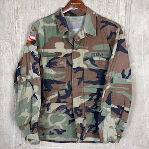 May include: Camouflage pattern military jacket with the words "U.S. ARMY" above the left chest pocket. The jacket has a button-down front, two chest pockets, and an American flag patch on the left sleeve.