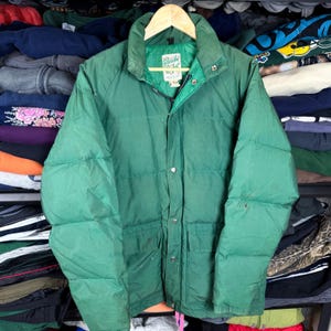 May include: A green, quilted winter jacket with a high collar and multiple pockets. The jacket has a zipper and snap closures. The label inside the collar reads "Colorado Jacket". The jacket is hanging on a wooden hanger.