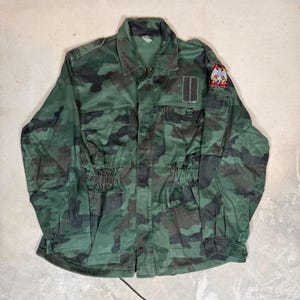 May include: A long-sleeve camouflage field jacket featuring a green, black, and brown pattern. The jacket includes a collar, a front button closure, four front pockets, reinforced elbow patches, and a small embroidered eagle patch on the left sleeve.