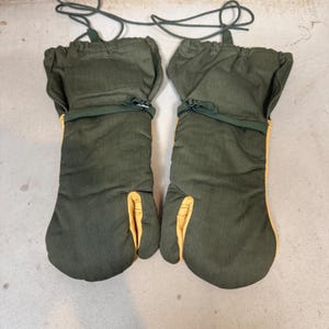 Puede incluir: Un par de manoplas de color verde oliva con detalles de cuero amarillo en el pulgar y los paneles laterales. Cada manopla cuenta con una correa ajustable de color verde oscuro en la muñeca y un cordón largo verde en la parte superior.