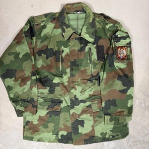 以下が含まれることがあります： 緑、茶色、黒の色合いのカモフラージュジャケット。 ジャケットは襟付き、複数のポケット、ジッパー開閉が特徴です。 上袖には紋章のパッチが見えます。 ジャケットは丈夫な生地で作られています。