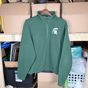 Puede incluir: Un jersey de forro polar verde con cuello alzado y cremallera de un cuarto de largo, con el logotipo blanco del casco espartano en el pecho. La parte superior de manga larga está colgada de una percha de madera.