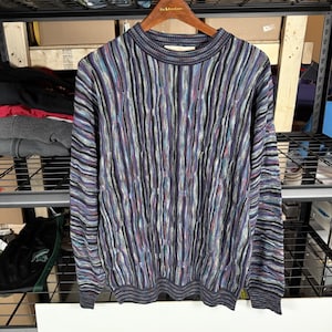 May include: A long-sleeved sweater with a ribbed pattern in shades of purple, blue, black, and white. The sweater is hanging on a wooden hanger. The ribbed design runs vertically down the garment.