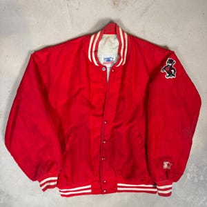 Puede incluir: Chaqueta roja Starter con rayas blancas en los puños y el dobladillo. La chaqueta tiene un cuello blanco con rayas rojas y un logotipo de Starter. Un parche con un personaje de dibujos animados está en la manga derecha.