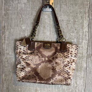 Peut inclure: Un sac cabas Coach avec un extérieur à motif peau de serpent marron et beige. Le sac comprend des garnitures en cuir marron foncé, des poignées en chaîne en métal doré et une petite plaque logo en métal doré sur le devant avec la marque Coach.