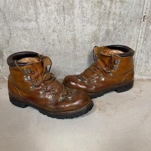 May include: A pair of well-worn, brown leather hiking boots. The boots have metal eyelets, thick laces, and a dark brown collar. The soles are black and show signs of wear. The boots are designed for outdoor activities.