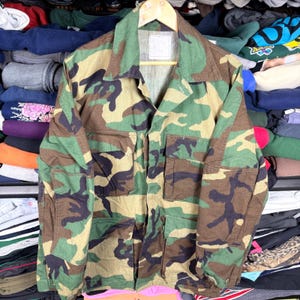 Könnte beinhalten: Eine Tarnjacke in Grün-, Braun- und Beigetönen. Die Jacke hat einen Kragen, vier Vordertaschen und einen Knopfverschluss. Sie hängt an einem Holzbügel.