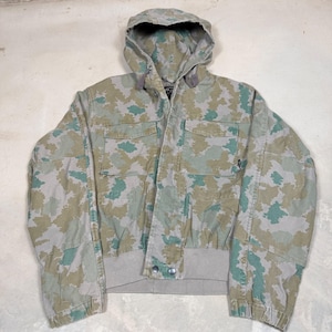 May include: A hooded camouflage jacket in shades of green, tan, and gray. The jacket features a front zipper closure, two chest pockets, and a ribbed hem and cuffs. The jacket has a classic military-inspired design.