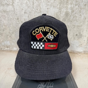 May include: A black corduroy baseball cap featuring an embroidered patch on the front. The patch displays the word CORVETTE in gold lettering above crossed red and checkered flags, with a black and white checkered pattern and a gold Chevrolet logo.
