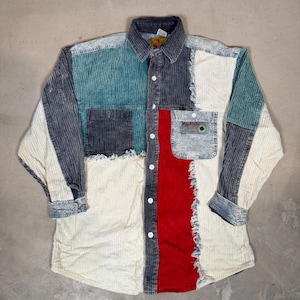 May include: A long-sleeved button-down shirt constructed from multi-coloured corduroy fabric panels. The shirt features a patchwork design in shades of cream, teal, charcoal grey, and red, with frayed edges and a single chest pocket with a label.