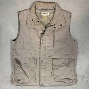 Puede incluir: Un chaleco acolchado de color beige con cuello, cremallera y cierres de botón a presión. El chaleco tiene dos bolsillos delanteros grandes con cierres de botón a presión y un bolsillo pequeño en el pecho. La etiqueta L.L.Bean es visible dentro del cuello.
