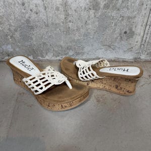 May include: A pair of wedge sandals featuring a brown cork-textured platform sole and a white crocheted strap design. The footbed is brown with a white heel patch displaying the brand name Muda in black text. The sandals have a thong-style toe post.