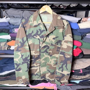 May include: Camouflage pattern jacket with a button-down front, two chest pockets, and two lower pockets. The jacket is made of a durable fabric and features a classic military-inspired design. The color palette includes shades of green, brown, and black.