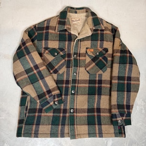 Puede incluir: Una camisa de manga larga con botones y un patrón de cuadros en tonos tostado, verde oscuro y azul marino. La camisa tiene un forro de estilo sherpa en color tostado, cuello en punta, dos bolsillos en el pecho con solapas y etiqueta Woolrich.