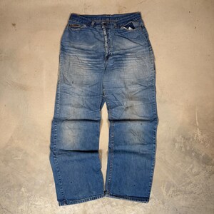 Può includere: Un paio di jeans blu a vita alta con chiusura a bottoni. Il tessuto presenta un lavaggio sbiadito con pieghe naturali sulla parte superiore delle cosce e sottili segni di usura lungo le gambe e sull'orlo.