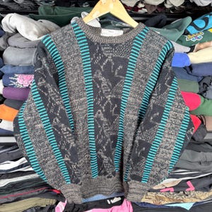 Op de afbeelding: Een vintage trui in tinten grijs, zwart en teal. De trui heeft verticale teal strepen en een patroon op de voorkant. Het wordt gepresenteerd op een houten hanger.
