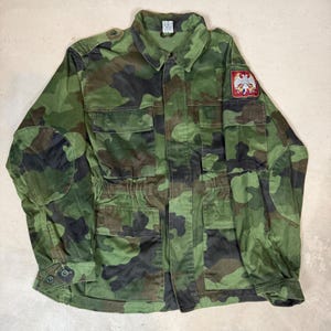 May include: A long-sleeve camouflage field jacket featuring a green, brown, and black pattern. The jacket includes a button-up front, four front pockets, shoulder epaulets, and elbow patches. A patch with a white eagle emblem is on the left sleeve.