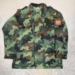 May include: A camouflage field jacket featuring a woodland pattern in shades of green, brown, and black. The jacket includes a fold-down collar, shoulder epaulets, multiple front pockets with zippers, and a patch on the left sleeve with an emblem.