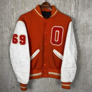 Könnte beinhalten: Vintage orange-weiße Collegejacke. Die Jacke hat weiße Ärmel mit der roten Nummer "69" und ein großes rotes "O" auf der Brust. Kragen und Manschetten haben orange-weiße Streifen. Die Jacke ist zugeknöpft und hängt an einem Holzkleiderbügel.