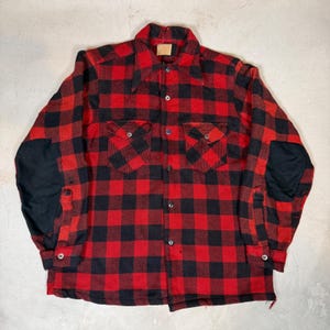 May include: A long-sleeve button-down shirt featuring a red and black buffalo plaid pattern. The shirt includes two chest pockets with buttoned flaps, a standard collar, and solid black fabric patches sewn onto the outer elbows of both sleeves.