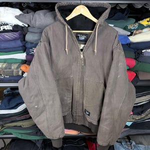 Puede incluir: Chaqueta con capucha Walls Blizzard marrón con cierre de cremallera, dos bolsillos delanteros y forro interior negro. La chaqueta está colgada de una percha de madera. El logotipo de Walls Blizzard es visible en la etiqueta.