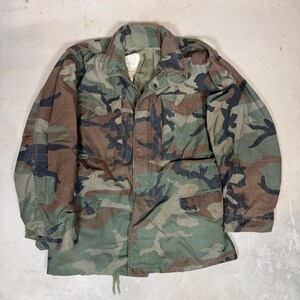 May include: A long-sleeve field jacket featuring a woodland camouflage pattern in shades of olive green, brown, and black. The jacket includes a fold-down collar, a front zipper closure, two chest pockets with flaps, and two lower pockets with flaps.