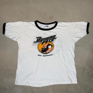 Puede incluir: Una camiseta blanca tipo ringer con ribetes negros en el cuello y las mangas. El frente presenta un gráfico con el texto Dynamo Fats sobre un círculo amarillo que contiene la ilustración de un hombre barbudo con gafas y el texto Says...Get Foozed!.