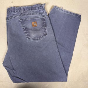 May include: Faded blue denim jeans with a classic five-pocket design. The jeans feature a brown leather patch on the back pocket with a logo. The jeans are folded and appear to be pre-worn.